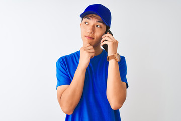 Wall Mural - Chinese deliveryman wearing cap talking on the smartphone over isolated white background serious face thinking about question, very confused idea