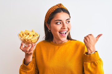 Poster - Young beautiful woman holding bowl with macaroni pasta over isolated white background pointing and showing with thumb up to the side with happy face smiling