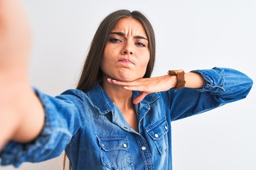 Beautiful woman wearing denim shirt make selfie by camera over isolated white background cutting throat with hand as knife, threaten aggression with furious violence