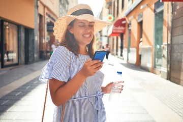 Wall Mural - Young beautiful woman smiling happy walking on city streets on a sunny day of summer using smartphone