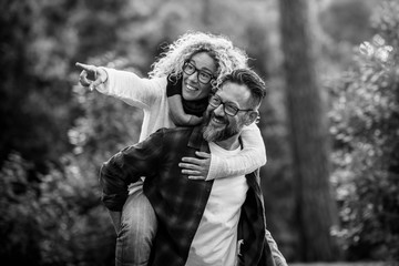 Wall Mural - Black and white image with happy and joyful adult caucasian couple having fun in outdoor forest leisure activity - man carry woman and both laugh a lot with love and friendship