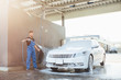 Professional bearded  washer in blue uniform washing luxury car with water gun on an open air car wash.