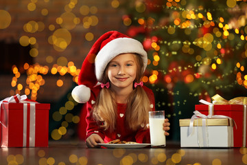 Canvas Print - Little girl in Santa Claus cap with cookies and milk near Christmas tree indoors