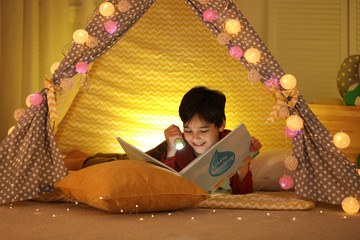 Canvas Print - Boy with flashlight reading book in play tent at home