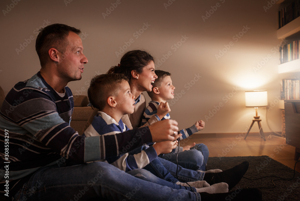 Parents with they children  sitting at home on pleasant evening and playing games on console.They challenge each other to win . - obrazy, fototapety, plakaty 