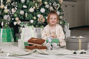 Merry Christmas and Happy Holidays! Beautiful little girl in a dress with a gift in her hand sitting near the Christmas tree. Winter holidays. Happy New Year. Portrait. Winter holidays concept