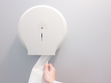Hand-pulling The Tissue Roll Out A White Box Mounted On A White Wall In A Public Restroom.