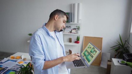 Wall Mural - Tilt up zoom out of printed application interface samples on office desk and young entrepreneur standing in office, looking at patterns for different devices on laptop screen and having phone talk