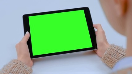 Over shoulder view - woman sitting at table and looking at black digital tablet computer device with blank green screen in cafe. Technology, chroma key, template, mock up and entertainment concept