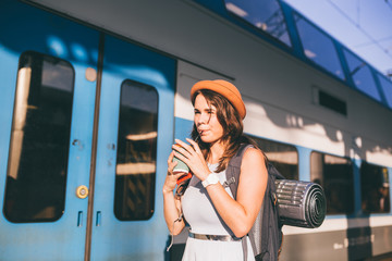 Tourism and travel in the summer. Vacations for the student. Work and travel. Caucasian young woman drinks coffee on the platform of the railway station against the background of the train