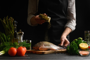 Wall Mural - Seafood, Chef cooks fish, sprinkles with spices, frozen in motion. On a black background with ingredients on the table. Healthy and healthy food, sale of seafood, fish shops