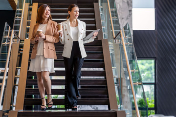 Wall Mural - Two asian businesswomen walking and talking during coffee break in modern office or coworking space, coffee break, relaxing and talking after working time, business and people partnership concept