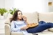 © Andrii - Tired woman is chewing pizza, while laying on the white sofa. She is watching TV shows, being on blurred background.