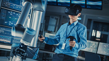 Professional Japanese Development Engineer In Blue Shirt Is Controlling A Futuristic Robotic Arm With A Virtual Reality Headset And Joysticks In A High Tech Research Laboratory With Modern Equipment.