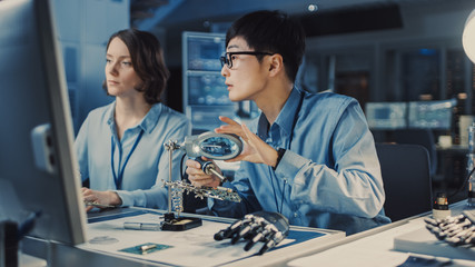 Japanese Development Engineer in Blue Shirt is Soldering a Circuit Board in a High Tech Research Laboratory with Modern Equipment. His Colleague Asks Him a Questio and Points on the Compter Screen.