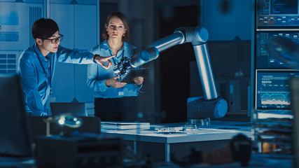 Leinwandbilder - Man and Woman Engineers with Laptop and a Tablet Analyse and Discuss How a Futuristic Robotic Arm Works and Moves a Metal Object. They are in a High Tech Research Laboratory with Modern Equipment.