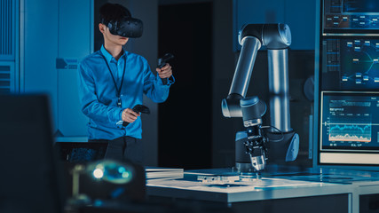 Leinwandbilder - Professional Japanese Development Engineer in Blue Shirt is Controlling a Futuristic Robotic Arm with a Virtual Reality Headset and Joysticks in a High Tech Research Laboratory with Modern Equipment.