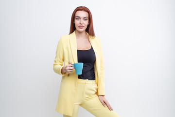 Wall Mural - A red haired girl in a yellow business suit holds a blue mug on a white background