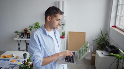 Wall Mural - Tilt up of printed app interface samples, papers and documents on office desk and young entrepreneur looking at patterns on laptop screen, then closing laptop and smiling at camera