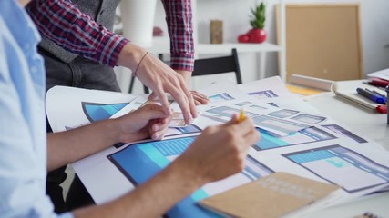 Wall Mural - Tilt down closeup of man explaining ideas to colleague at desk in office. Two web designers or marketing professionals discussing website interface samples for different devices printed on paper
