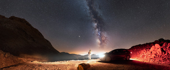 Panorama of milky way and two people standing near car and looking up to the sky