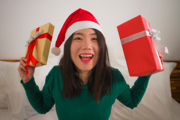 Christmas lifestyle portrait of young beautiful and happy Asian Chinese woman on bed in Santa Claus hat holding xmas presents and gift boxes excited