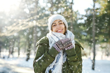 Winter portrait of funny smiling woman in woolen hat and long warm scarf in snowy winter park at frozzy sunny day. Flying snowflakes. Happy winter time, having fun