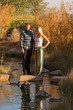 © Melani - Vertical image of a young couple standing on a stepping stone in a small stream with autumn foliage behind them watching some ducks swim in the water.