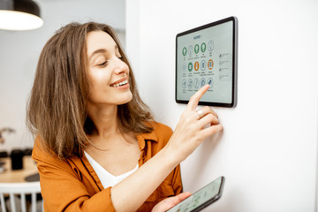 Young woman controlling home with a digital touch screen panel installed on the wall in the living room. Concept of a smart home and mobile application for managing smart devices at home