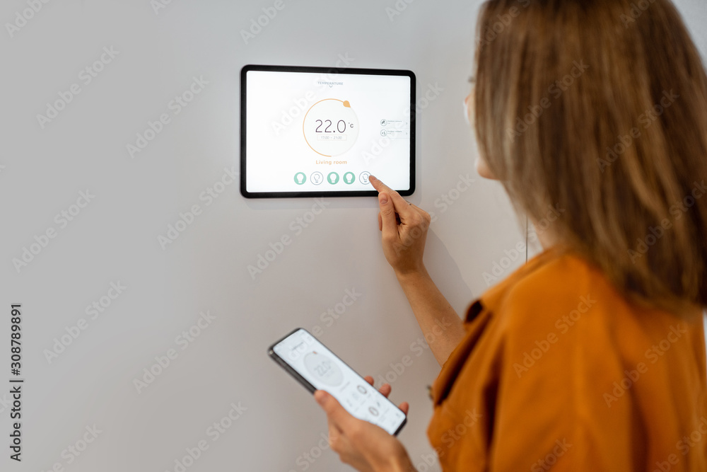 Young woman controlling temperature in the living room with smart phone and digital touch screen panel. Concept of heating control in a smart home - obrazy, fototapety, plakaty 