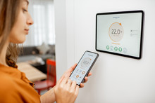 Young Woman Controlling Temperature In The Living Room With Smart Phone And Digital Touch Screen Panel. Concept Of Heating Control In A Smart Home