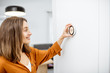 © rh2010 - Young woman regulating heating temperature with a modern wireless thermostat installed on the white wall at home. Smart home heating regulation concept