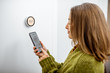 Woman dressed in green sweater regulating heating temperature with a modern wireless thermostat and smart phone at home. Synchronization of thermostat with mobile devices concept