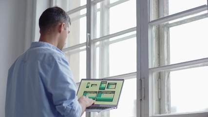 Wall Mural - Side view of young designer creating application interface for different devices on laptop and looking through window thoughtfully