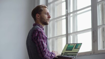 Wall Mural - Side view of young web designer in eyeglasses examining application interface samples for different devices on laptop and looking through window thoughtfully