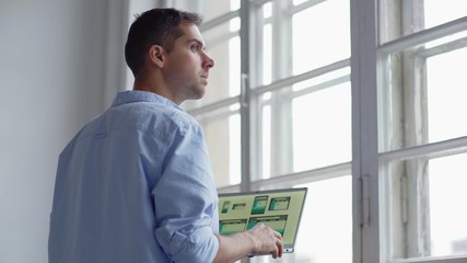 Wall Mural - Side view of young web designer looking through window thoughtfully while working on application interface design on laptop computer
