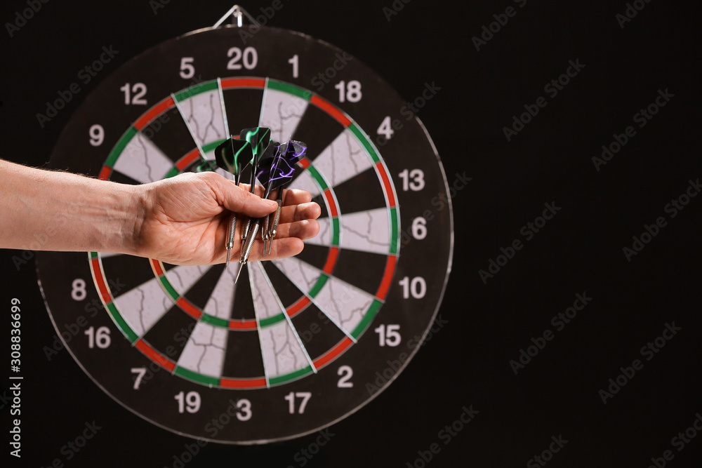 Male hand with darts and dartboard on dark background