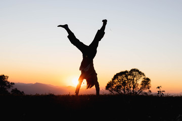 Wall Mural - Silhouette happy child playing upside down outdoors in summer park walking on hands at sunset