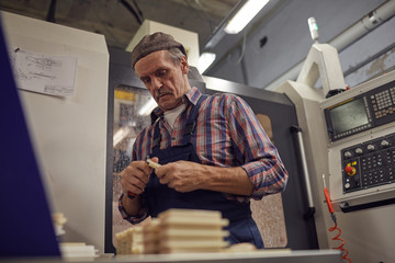 Senior man in work wear cutting details with instrument while standing near the lathe in the factory