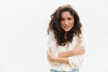 Wall Mural - Desperate frustrated woman feeling cold, trembling, hugging herself for warm. Wavy haired young woman in casual shirt standing isolated over white background. Fever or freezing concept