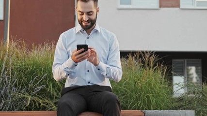 Wall Mural - Joyful bearded businessman using smartphone while sitting on bench outdoors