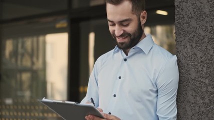 Wall Mural - Cheerful bearded businessman checking something in clipboard and becoming happy outdoors