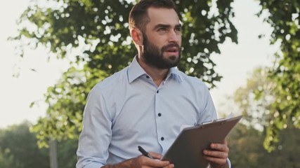 Wall Mural - Serious bearded businessman checking something in clipboard and becoming unhappy while sitting in park outdoors