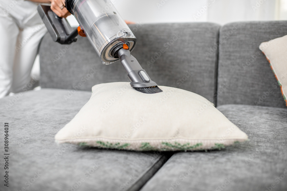Woman cleaning sofa with a modern cordless vacuum cleaner with a special nozzle, close-up. Dry cleaning concept with wireless vacuum cleaner - obrazy, fototapety, plakaty 