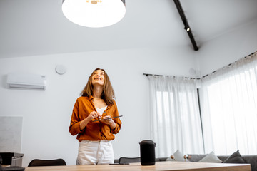 Wall Mural - Young woman controlling home light with asmart home in the living room. Concept of a smart home and light control with mobile devices