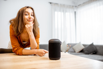 Wall Mural - Portrait of a young and cheerful woman with a smart wireless column at home. Concept of smart home and voice command control