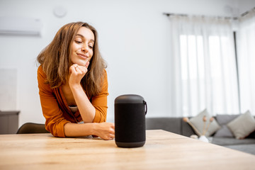 Wall Mural - Portrait of a young and cheerful woman with a smart wireless column and phone at home. Concept of smart home and voice command control