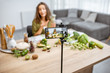 © rh2010 - Young woman recording on a smart phone her vlog about healthy eating. Sitting at the table with lots of green vegan food ingredients at home