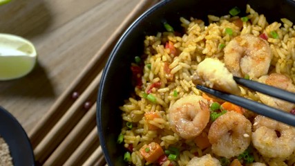 Wall Mural - Quick and healthy Asian meal made of fried rice, fresh shrimps, lime and vegetables in a black bowl isolated on white background. Someone’s hand takes a shrimp with chopsticks. 