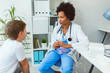 © lordn - Smiling female afro american doctor general practitioner talks and amuses child before medical examination